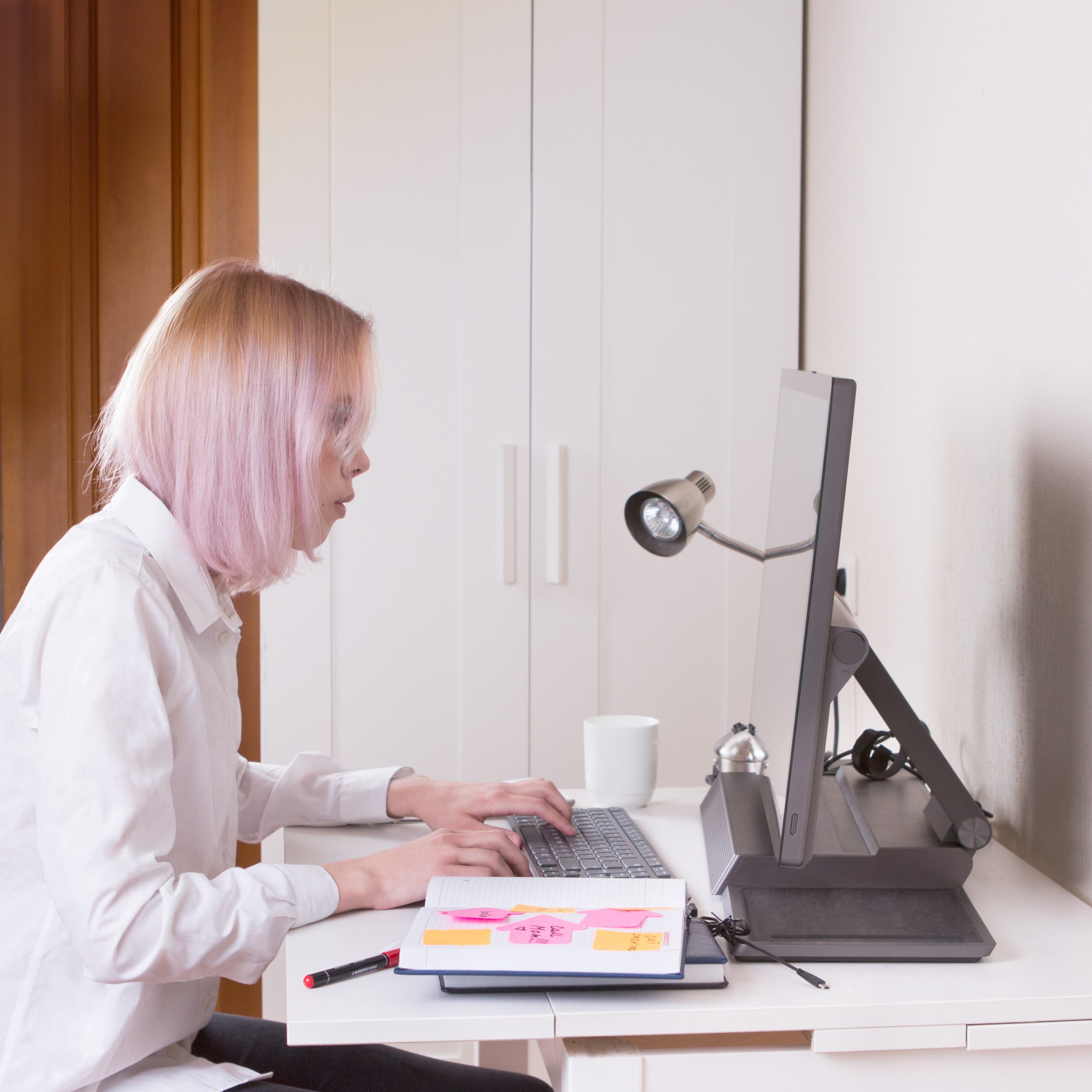 Young girl with pink hair is working at home office on computer, using her all-in-one (AIO PC) desktop.
